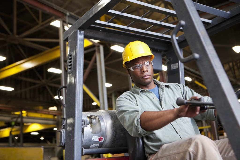 Male warehouse worker wearing a hard hat and safety goggles while driving a forklift.